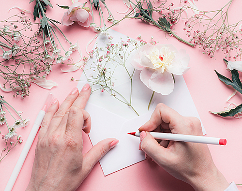 Female Hands write on envelope with flowers on pink tables background.  Wedding, invitation ,Valentine day,  Mother's Day greeting and love concept. Flat lay, top view, copyspace