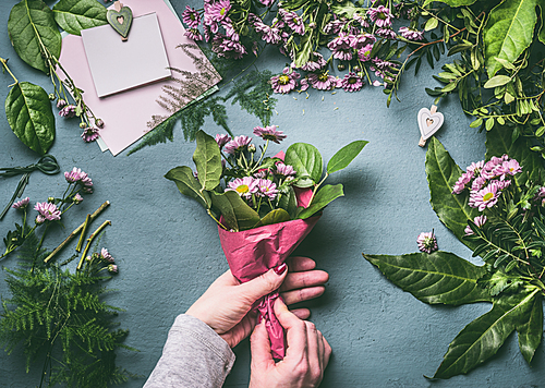 Female hands making lovely bouquet of pink flowers with  wrapping paper  on florist workspace, top view. Florist work, step by step