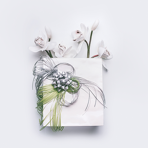 Paper gift bag with green ribbons and orchid flowers on white desktop background, top view