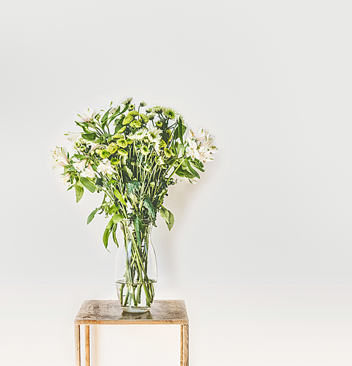 Beautiful green flowers bunch with falling petals in glass vase at white wall background. Home interior and decor ideas