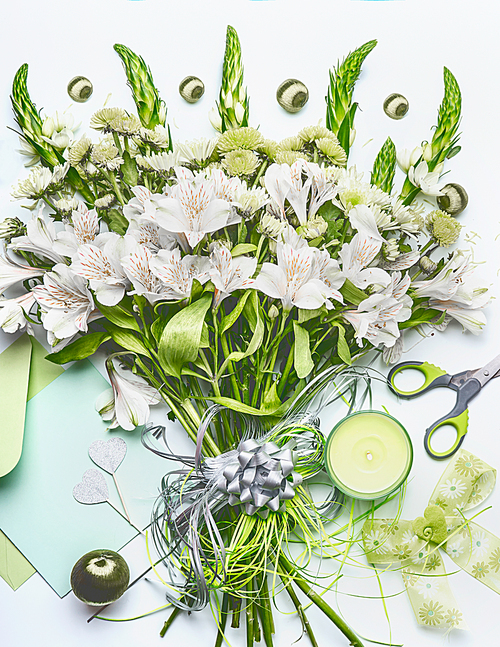 Beautiful festive bouquet  with green flowers on white desk background with envelope and paper card mock up, ribbon, candle and scissors, top view. Holidays greeting concept, flat lay