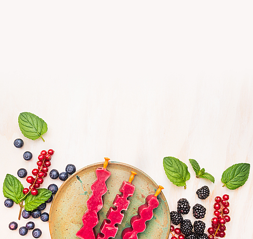 Ice cream pops in plate with summer berries:  red currant, blackberries, blueberries and peppermint leaves on white wooden background, top view place for text, frame
