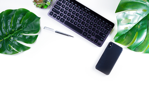 Flat lay home office workspace with black keyboard and green monstera leaves on white background