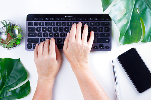 Flat lay home office workspace with someones hands typing on black modern keyboard and green leaves on white background