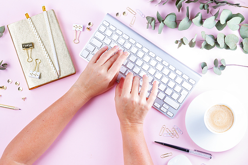 Flat lay home office workspace -someone hands typing on white modern keyboard on pink background with copy space