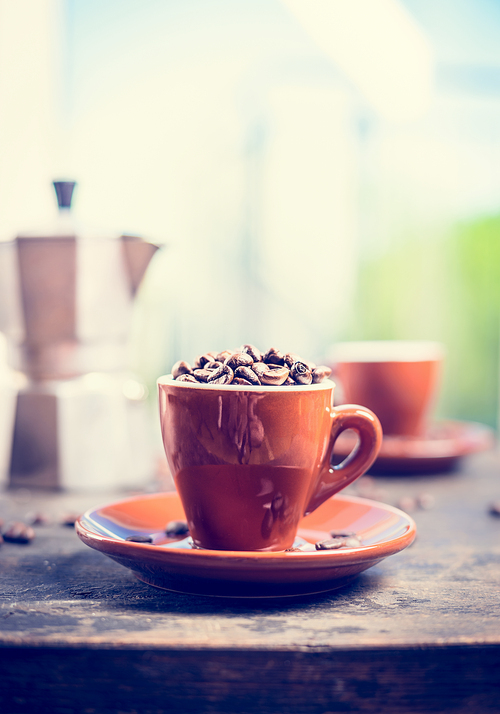 Brown espresso cup full of coffee beans on kitchen table with coffee pot on nature background