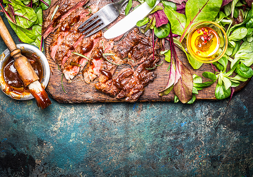 Sliced grilled steak served with green salad, Barbecue sauce and cutlery on wooden gutting board and rustic background, top view, border