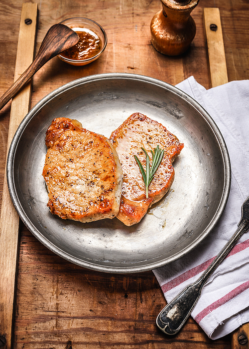 Two roasted pork steaks on rustic metal plate on wooden background, close up