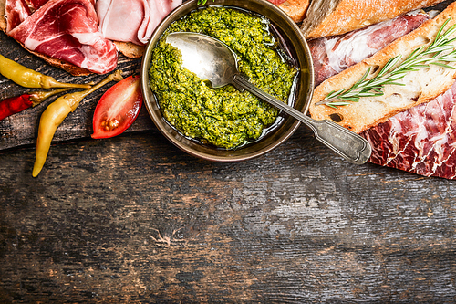 Green pesto and meat plate with bread and antipasti on rustic wooden background, top view, border. Italian food concept
