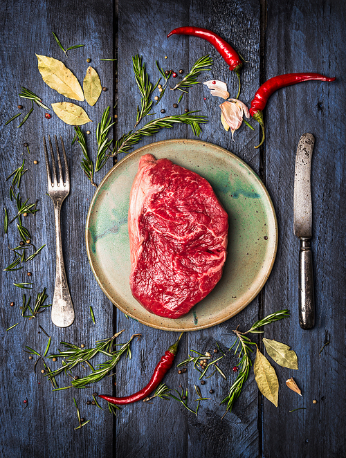 Raw Striploin Steak on  plate with  knife and fork to lay with herbs and spices, blue wooden background, top view