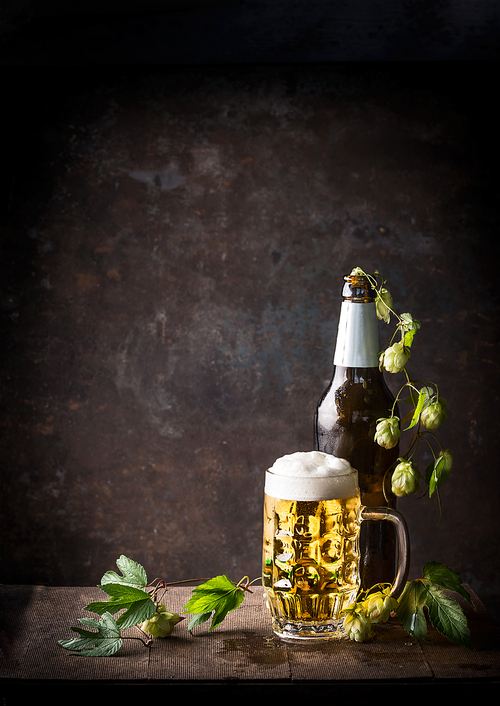 Glass bottles and mug of beer with cap of foam and hops on table at dark rustic background, front view, Still life