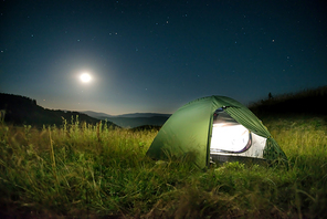 Illuminated green tent in the mountains at night under dark sky with many stars