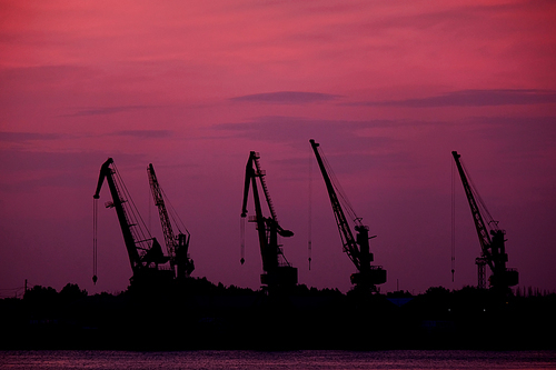 Five Silhouettes Of Wharf Cranes In Front Of Pink Sunset Sky With Violet Clouds.