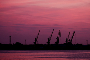 Many Silhouettes Of Wharf Cranes In Front Of Pink Sunset Sky With Violet Clouds. A lot of space for text.