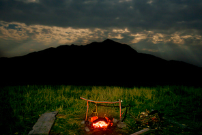 Campfire on green lawn near mountain at night with dark sky and moon light