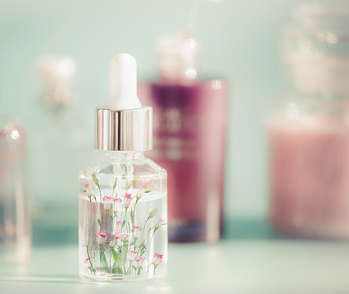 Transparent cosmetic bottle with liquid, pipette and little pink flowers. Floral essence or herbal extract serum on table, front view. Beauty and modern skin care concept