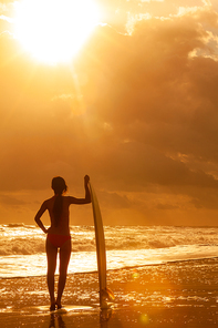 rear view of beautiful  young woman surfer woman in pink bikini with white surfboard on a beach at sunset or sunrise