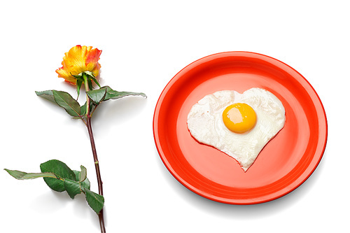 Heart shaped egg on the red plate next to fresh rose.