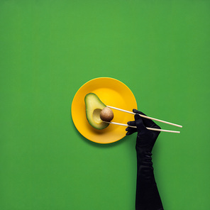 Creative concept photo of avocado with hand and chopsticks on painted plate on green background.