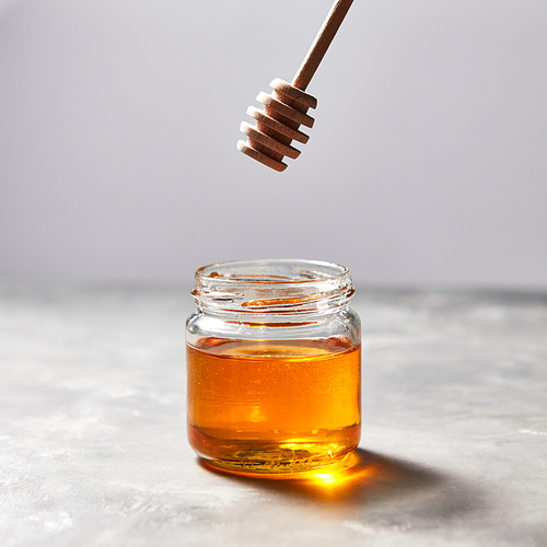 Wooden stick above a glass jar with aromatic natural organic honey on a gray concrete table, place for text. Jewish New Year healthy holiday concept. Traditional useful sweetness.