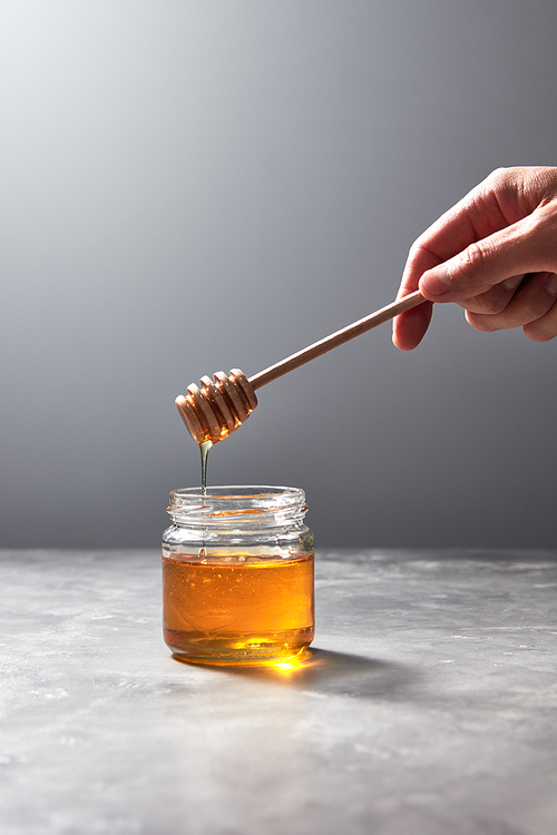 Woman hand hold a wooden stick with dripping flower honey into a glass jar on a gray background, place for text. Pure organic sweet goodness.