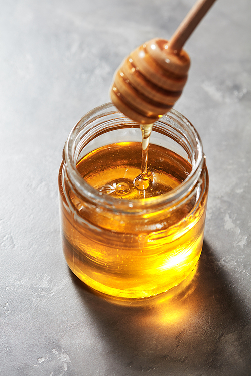 Dripping sweet natural organic honey from stick into glass jar on a gray marble background, pure natural raw sweet goodness.