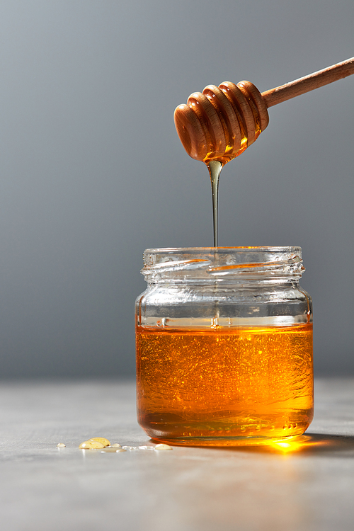 Wooden dipper with drains raw organic honey into a glass full jar with sweet syrup on a gray marble background, pure natural sweet goodness.