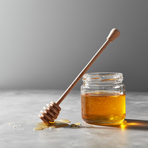 Wooden dipper in a puddle of syrup and a glass jar with fresh natural organic honey on a gray concrete background, place for text. Jewish New Year holiday concept.