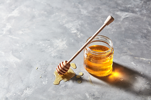 Sweet floral natural honey in a jar and wooden spoon in a puddle of syrup on a gray marble background, copy space. Jewish rosh hashanah holiday concept.