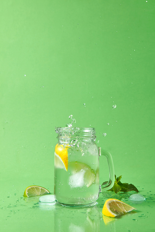 Splash and drops of lemonade from a glass and slices lime, lemon, mint sprig on a green table. Glass jar with homemade sparkling lemonade on green background with copy space.