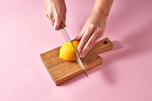 Citrus fruits on cutting board. Female hands cutting a yellow ripe lemon on half on a wooden board on a pink background. Concept vegetarian food.