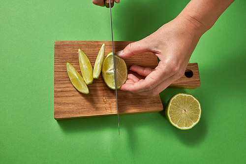 Top view of female cutting a half of green ripe lime with slices on a wooden cutting board on a green background. A sharp knife in her hands.