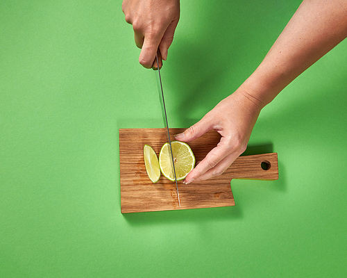 Slices of natural organic green lime, cutting by female hands on a wooden board on green background. Concept of healthy food.