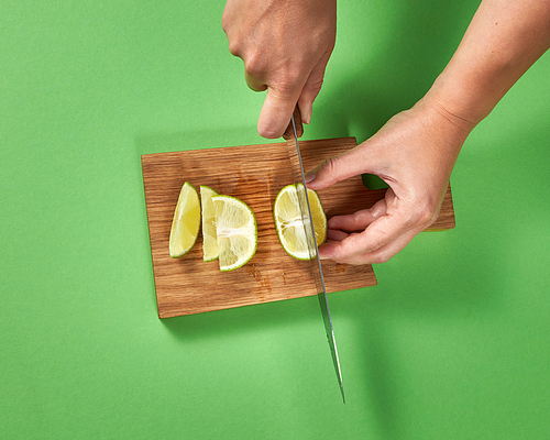 Top view of girl cutting a half of organic ripe lime with slices on a brown board on a green background. A sharp knife in her hands.