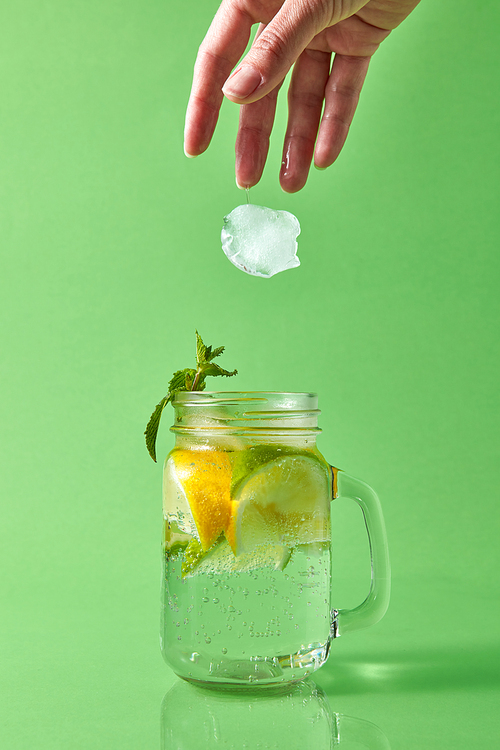 Glass jar on a green with homemade sparkling cocktail with slices of citrus fruits, green leaves of mint. A woman's hand throws a piece of ice into a glass jar.