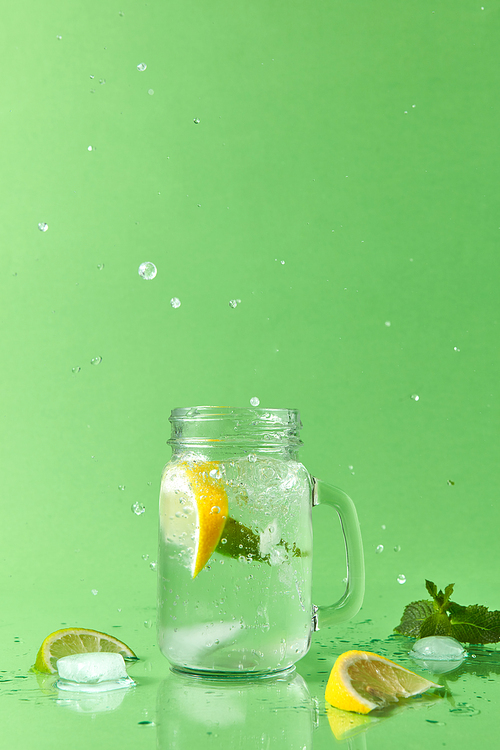 Glass jar on a green table with homemade sparkling lemonade with slices of lime and lemon, green leaves of mint. Splash lemonade from a glass and slices lime, lemon on a green table .