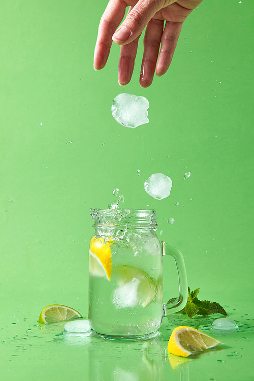Splashes and drops of a natural cocktail scatter on the surface of the table. A female hand throws ice cubes into a glass with a homemade natural cocktail on a green background.