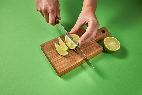 Wooman hands cut a juicy green ripe lime to slices with knife on a wooden cutting board on a green background with copy space. Concept of healthy food.