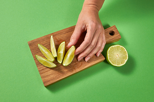 Top view of citrus background. Slices of lime on a board on a green table. A female hand puts slice of ripe fresh green lime on a wooden cutting board with copy space.