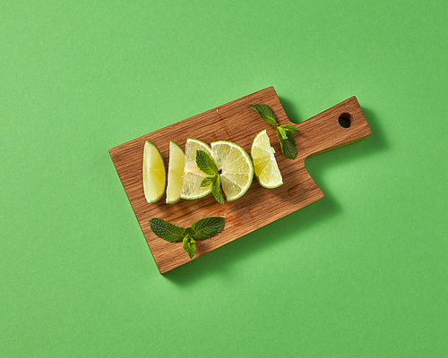 Top view of citrus fruits green lime with sprigs of green mint on brown board on a green background. Concept of cold alcoholic or non-alcoholic summer drinks.