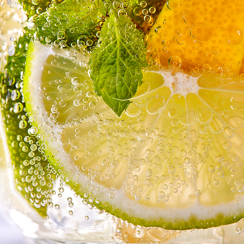 Green leaf of mint, pieces of lemon and lime with bubbles in a transparent glass. Macro photo of summer cold mojito