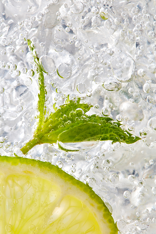 Macro photo of a piece of lime and mint leaf in a glass with ice and bubbles. Cold summer drink concept