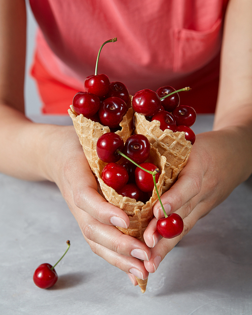 Three sweet waffle cones with ripe cherries holding girls hand on a gray background. Summer concept of homemade desserts.