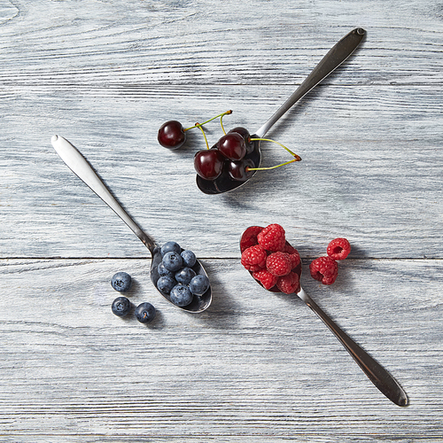 Red ripe juicy berries - cherry and raspberry, blueberry in the three metal spoons and near them on a gray wooden background. Top view