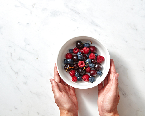 Tasty berries, freshly picked fruits on ceramic plate in the female hands on a concrete background. Concept of healthy food with copy space. Flat lay