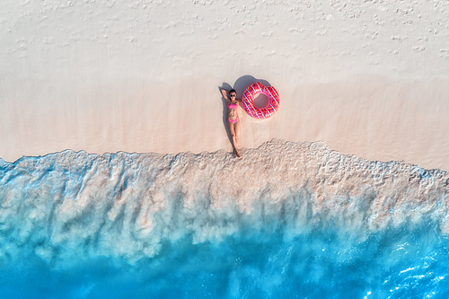 Aerial view of the beautiful young lying woman with pink donut swim ring on the white sandy beach near sea with waves at sunset. Summer holiday. Top view of slim girl, clear azure water. Indian Ocean