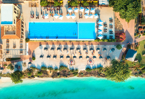 Aerial view of pool, umbrellas, sandy beach with green trees, sea. Indian ocean at sunset in summer. Zanzibar, Africa. Top view. Landscape with hotel, azure water, parasols, palm trees. Luxury resort