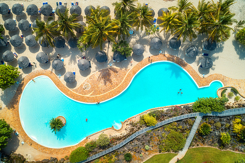 Aerial view of pool, umbrellas, sandy beach with green trees. Coast of Indian ocean at sunset in summer. Zanzibar, Africa. Top view. Landscape with azure water, parasols, palm trees. Luxury resort