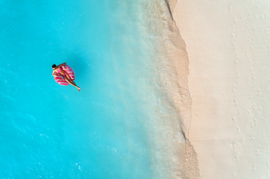 Aerial view of a young woman swimming with the donut swim ring in the clear blue sea with waves at sunset in summer. Tropical aerial landscape with girl, azure water, sandy beach. Top view. Travel
