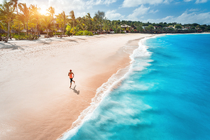 Aerial view of the running young woman on the white sandy beach near sea with waves at sunset. Summer holiday. Top view of sporty slim girl, clear azure water. Indian Ocean. Lifestyle and sport